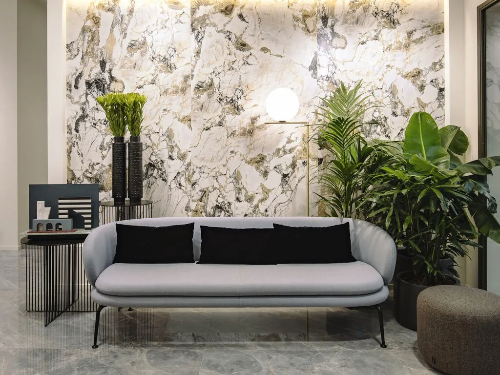 The image shows a stylish, contemporary living room featuring high gloss Alpine White granite effect wall panels as the backdrop. The wall panels have a striking pattern with white, black, and gold veining, giving the space a luxurious and natural feel. In front of the wall is a light gray modern sofa with black accent pillows, surrounded by a variety of plants that add a refreshing touch of greenery. A sleek side table with decorative elements and a floor lamp with a spherical light enhance the room's minimalist and cozy ambiance. The marble flooring ties the design together with a polished, reflective surface.