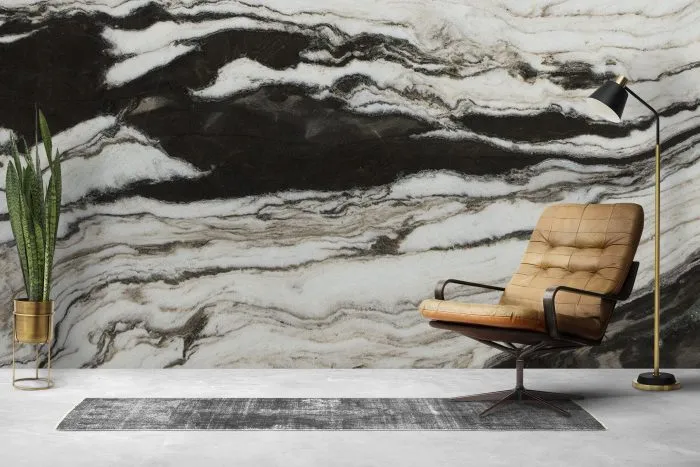 The image features a room with walls adorned with Calacatta Black Vein Marble Effect Wall Panels, which have a high gloss finish. The marble effect is characterized by a dark background with contrasting white and grey swirling patterns that mimic natural marble veining. In the foreground, there is a modern, tan leather chair with a metal base positioned on top of a textured grey area rug. To the left of the chair stands a tall floor lamp with a slim profile and an angled light source, casting light towards the chair. Adjacent to it is a small golden pot holding a green potted plant that adds a touch of color to the scene. The overall aesthetic suggests an elegant and contemporary interior design.