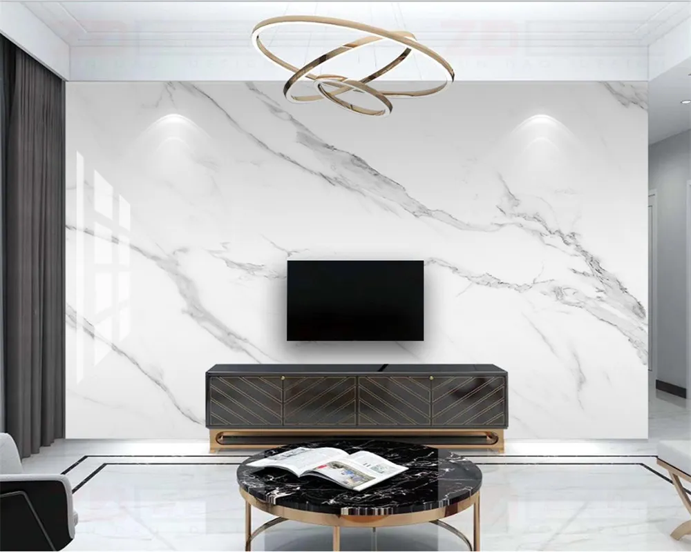 The image shows a modern, elegant living room with a wall-mounted television. The backdrop features high gloss Capraia Bianco marble effect wall panels. The marble design has gray veining that contrasts with the white background, creating a luxurious and contemporary look. Below the TV, there is a dark-colored low console with gold-accented lines. In front of the console, a black marble coffee table complements the minimalist and sophisticated aesthetic of the space. Wall lights and a circular pendant light add a touch of modernity.